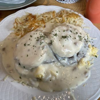AVGO - The Outlaw Biscuits & Gravy - A white textured plate contains biscuits smothered in white gravy with two eggs on top, garnished with herbs. To the side, there are hash browns. The background shows a table with a pink mug, a basket of bread, and other dining items, indicating a casual dining environment. - Breakfast & Brunch Near Me - Saint John, Indiana