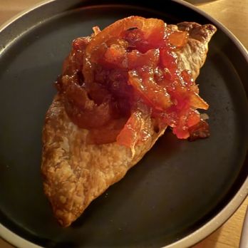 Baleia - Duck liver empada - A fried empanada with a crispy exterior is served on a black plate, topped with a tomato-based sauce containing diced tomatoes and onions. - Wine Bars Near Me - Boston, Massachusetts