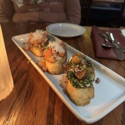 Vinaio Bruschetta - Three pieces of bruschetta with various toppings, including diced tomatoes, herbs, cheese, and nuts, are served on a white rectangular plate. The plate is placed on a wooden table in a restaurant, with a person seated in the background, a small empty plate, and utensils wrapped in a napkin nearby.