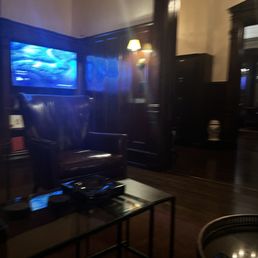 The image shows a lounge area with dark wood paneling and trim. A leather armchair is positioned near a small glass-top table, which holds electronic devices. A television mounted on the wall displays a blue scene, and a wall-mounted lamp provides warm lighting. The space appears cozy and traditional, with a focus on comfort and subdued lighting.