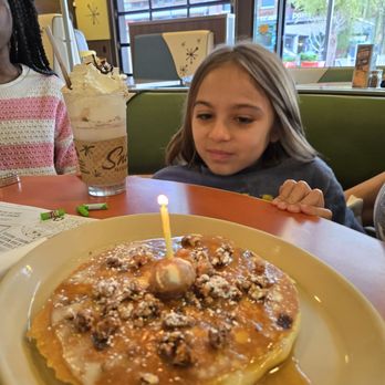 Snooze, an A.M. Eatery - A girl sits at a table in a restaurant, gazing at a birthday dessert with a lit candle, alongside a milkshake topped with whipped cream. - Breakfast & Brunch Near Me - Gilbert, Arizona