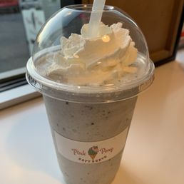 Black sesame shake - Cookies and cream milkshake with whipped cream topping in a clear plastic cup, placed on a white surface near a window.