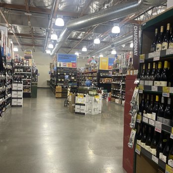 BevMo! - The image shows the interior of a liquor store with multiple aisles lined with shelves stocked with wine bottles. Bright overhead lighting illuminates the space, and promotional signs are visible above the shelves. The store has an industrial ceiling with exposed ductwork and a polished concrete floor. There are various displays and signs indicating sales and product information. - Beer, Wine & Spirits Near Me - Pasadena, California