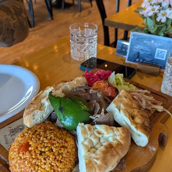Meat Point - Meat doner with bulgur - A meal consisting of a doner wrap filled with meat, vegetables, and sauce, served alongside a portion of bulgur on a wooden serving board. The restaurant interior includes tables with chairs, a glass of water, and a flower arrangement on the table. The background shows a casual dining environment with additional tables and chairs, and a display case or refrigerator. - Turkish Near Me - Toronto, Ontario