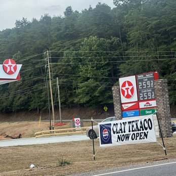 Clay Texaco - View from road - The image shows a Texaco gas station sign with fuel prices listed for regular, credit/debit, and diesel. In front of the sign, a banner reads 'Clay Texaco Now Open' with Pepsi branding. The background includes a wooded hillside, utility poles, and a parked car. The scene is outdoors along a roadside with a partly cloudy sky. - Convenience Stores Near Me - Birmingham, Alabama
