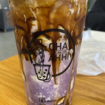 Cha’ahh - Ube Macapuno Crème Brûlée - A tall cup from Cha’ahh bubble tea café contains a purple and white beverage with chocolate syrup drizzled on top, served on a wooden table. - Bubble Tea Near Me - Gallup, New Mexico