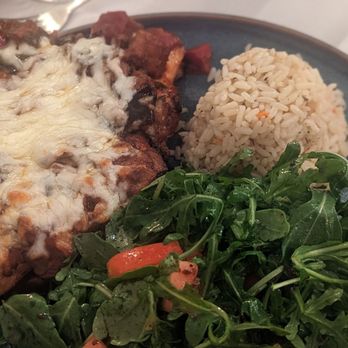 Athos - A plate contains a piece of baked chicken topped with melted cheese, accompanied by a portion of rice and a side salad with leafy greens and sliced tomatoes. The dish is presented on a dark ceramic plate, with a white tablecloth underneath. - Greek Near Me - Albany, New York