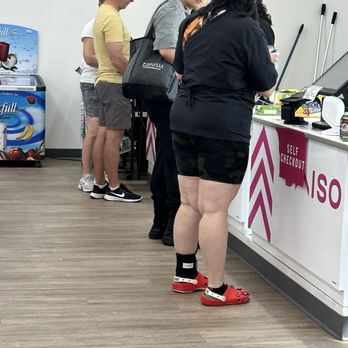 Daiso - Four people are standing in a line at the self-checkout counter of a retail store. The first person is a woman with black hair, wearing a black jacket and shorts, with red Crocs. The second person is a woman with glasses, wearing a gray shirt and black pants. The third person is a man in a yellow T-shirt, gray shorts, and a cap. The fourth person is a woman in a white shirt and shorts. The store has a white and pink color scheme, with a self-checkout sign and a cash register visible. - Department Stores Near Me - Mesa, Arizona