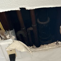 The slab leak! - A drywall opening exposes plumbing pipes and a water meter, with an electrical outlet and cord nearby.