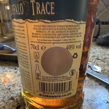 Lighthouse Wine & Spirits - Gold sticker covering the UK label. - A clear glass bottle of Buffalo Trace Kentucky Straight Bourbon Whiskey is positioned on a speckled kitchen countertop. The label is partially torn, obscuring some details, but the visible text indicates the bottle contains 70cl of whiskey with 40% alcohol by volume. The label includes information about the distillery, distribution, and a barcode. The background shows a knife and other kitchen items, suggesting a home setting. - Beer, Wine & Spirits Near Me - Beverly, Massachusetts