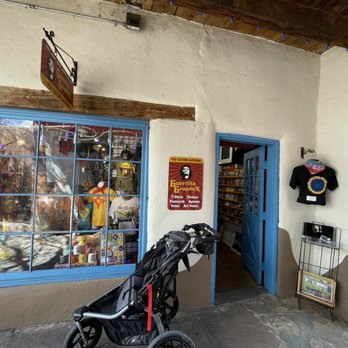 Guerrilla Graphix - The image shows the storefront of Guerrilla Graphix in Albuquerque, NM, with a large display window filled with various merchandise including T-shirts, stickers, and art prints. The window has a blue frame, and the entrance door is also painted blue. To the right of the door, there is a small display stand with additional items and a T-shirt hanging on the wall. A stroller is parked in front of the store, and the building has a rustic appearance with a wooden beam above the window and a textured stone floor. A sign with the store's name and offerings is visible near the entrance. - Printing Services Near Me - Albuquerque, New Mexico