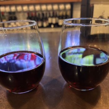 Timber Pizza - McLean - Glasses of pino noir - Two glasses of red wine are placed on a wooden table in a restaurant or bar, with a blurred background of bottles and decor. - Pizza Near Me - McLean, Virginia