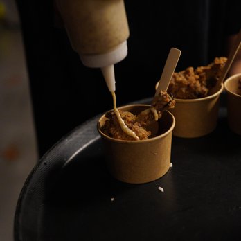 Ghost Chicken - Fried chicken pieces are placed in small paper cups on a black tray, with a person squeezing sauce from a bottle onto one of the pieces. - Comfort Food Near Me - Toronto, Ontario