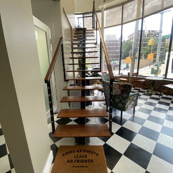 Artizen - Upper area for non membership working customers - The image shows a cafe interior with a spiral staircase that has wooden steps and a metal railing, positioned near large windows that let in natural light. The floor has a black and white checkered pattern, and there are chairs and tables visible near the windows. A doormat with the words "COME AS GUESTS LEAVE AS FRIENDS" is placed at the base of the staircase. - Coffee & Tea Near Me - The Bronx, New York