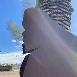 Sasquatcho, basking in the sun - A silhouette of a woman's face profile cutout is situated outdoors, with a large tree trunk and green foliage in the background. The sky is clear and blue, with sunlight causing a lens flare across the image. The cutout appears to be made of wood or similar material, painted in a dark color, and is mounted upright. The scene suggests a park or outdoor recreational area.