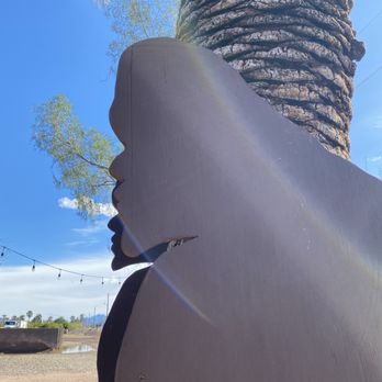Sasquatcho Taco - Sasquatcho, basking in the sun - A silhouette of a woman's face profile cutout is situated outdoors, with a large tree trunk and green foliage in the background. The sky is clear and blue, with sunlight causing a lens flare across the image. The cutout appears to be made of wood or similar material, painted in a dark color, and is mounted upright. The scene suggests a park or outdoor recreational area. - Mexican Near Me - Mesa, Arizona