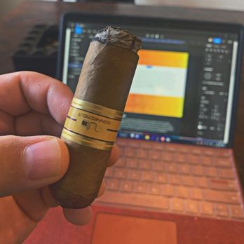 Padres Cigars - Lit cigar with laptop in background. - A person holds a cigar with a gold band in front of a laptop displaying a colorful document or webpage. - Cigar Bars Near Me - Corrales, New Mexico