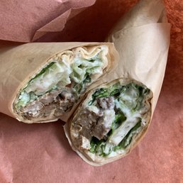 Lamb Gyro Sandwich - A sandwich wrap, sliced in half, displays layers of lettuce, cooked meat, and a creamy white sauce inside a soft flatbread. The wrap is partially unwrapped, showing the filling, and rests on a brown textured surface with a piece of parchment paper nearby.