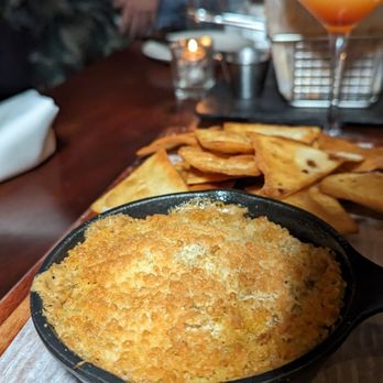 Tavern 157 - A baked casserole with a golden, crispy breadcrumb topping is served in a black dish, accompanied by fried chips and a cocktail in the background. - Lounges Near Me - Flushing, New York