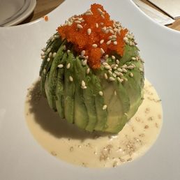 A culinary presentation of an avocado, sliced and arranged on a white plate, topped with orange fish roe and sprinkled with sesame seeds. The avocado is surrounded by a light-colored creamy sauce with sesame seeds, and the dish is served on a modern, curved white plate. The setting appears to be in a restaurant environment.