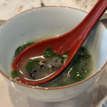Lily’s Kitchen - A bowl containing clear soup with green herbs and a red spoon, set on a light-colored surface. - Chinese Near Me - Malvern, Pennsylvania