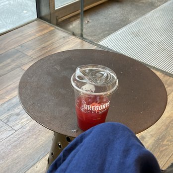 Gregorys Coffee - Strawberry Lemonade Refresher - A clear plastic cup containing a red strawberry lemonade drink with ice is positioned on a small, round, dark-colored table. The cup has a plastic lid with a straw hole and the Gregory's Coffee logo printed on it. The setting appears to be inside a coffee shop near a glass door, with wooden flooring and a metal threshold visible in the background. - Coffee & Tea Near Me - Washington, DC, District of Columbia