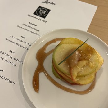 Leader - A dessert consisting of sliced apple and pear stacked in a cylindrical shape, garnished with a green chive, and topped with a dollop of fruit compote. The plate has a sauce drizzled around the fruit. A menu with the date September 27, 2022, and various dishes is visible in the background, indicating a dining setting at a restaurant. - Scandinavian Near Me - Cambridge, Minnesota