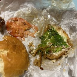 Blue Cheese Burger - The image shows a partially eaten burger on crumpled white paper. The burger bun is visible, with some lettuce, tomato slices, and pickles. The paper appears greasy, indicating the burger has been eaten. The background includes a wooden surface and a light blue surface. The food presentation suggests a casual dining or takeout setting.