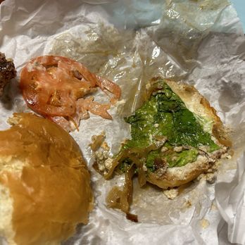 Fusion Burger - Heavenly blue cheese burger . Nothing but grease - The image shows a partially eaten burger on crumpled white paper. The burger bun is visible, with some lettuce, tomato slices, and pickles. The paper appears greasy, indicating the burger has been eaten. The background includes a wooden surface and a light blue surface. The food presentation suggests a casual dining or takeout setting. - Burgers Near Me - Eugene, Oregon