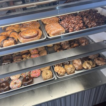 Donut Pantry - A refrigerated display case contains an assortment of donuts, including glazed, chocolate, sprinkled, and cinnamon varieties, along with other baked treats. - Donuts Near Me - Fallbrook, California