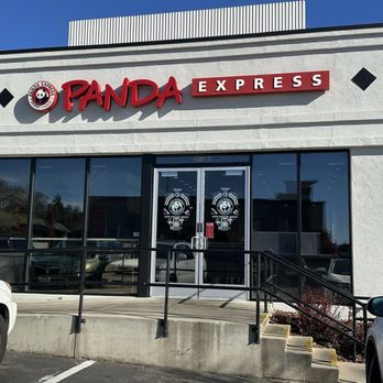 Panda Express - Outside - The photo depicts the exterior of a Panda Express restaurant, featuring a white facade with the Panda Express logo and signage prominently displayed. The entrance has double glass doors with Panda branding, and large glass windows on either side. There is a small ramp and stairs leading to the entrance, with parking spaces and vehicles visible in the foreground. The sky is clear and blue, indicating a sunny day. - Chinese Near Me - Sonora, California