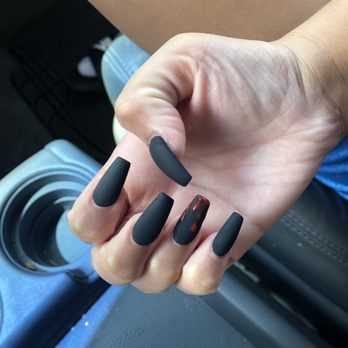 Cnn Nails - no-chip full set acrylic, black matte & "blood" design on one nail - A hand with long, matte black acrylic nails is shown, with one nail decorated with a blood-red design. The hand is positioned over a car interior, near a cup holder and part of a person's arm and leg are visible. The nails are well-manicured and the design adds a striking contrast to the matte black finish. - Nail Salons Near Me - Chicago, Illinois