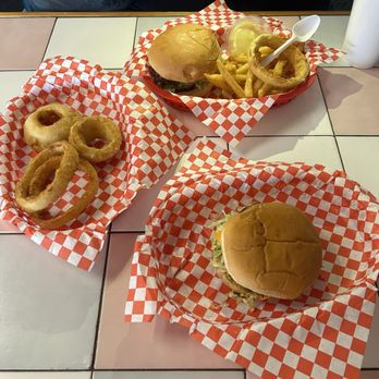 Rex's Hamburgers - Green Chile Cheeseburger Platter - The image shows two cheeseburgers with buns, lettuce, and cheese, placed on checkered paper on a tiled table. One burger is in a basket with fries and onion rings, with a small container of dipping sauce and a spoon. The other burger is on a separate piece of checkered paper. A person wearing a black jacket and white shirt is partially visible at the top of the image, and a white cup is on the right side of the table. - Burgers Near Me - Albuquerque, New Mexico
