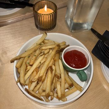 Nafsi - Truffle fries - French fries with ketchup are served on a white plate at a restaurant table, accompanied by a glass of water and a lit candle in a decorative holder. - New American Near Me - Chicago, Illinois