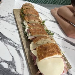 Sampler - corner bread fell off as he walked it over to our table. He toasted a new piece and brought it later. - A rectangular wooden board on a marble table holds several sandwiches with toasted bread, cheese slices, meats, and greens. The sandwiches are cut into halves or quarters, revealing fillings such as cheese, ham, and vegetables. The setting appears to be a restaurant or cafe, with a person sitting nearby and a patterned green wall in the background.