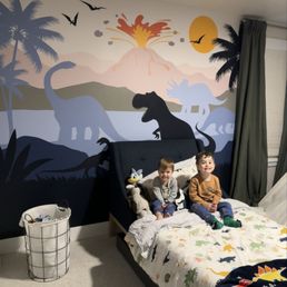 Dinosaur wallpaper and 2 little boys sitting on a twin sized bed