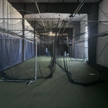 Adventure Commons - Upper Mezzanine with batting cages and pitching/catching turf - Indoor sports facility featuring batting cages with black nets, turf flooring, and supporting equipment such as a pitching machine and barriers. The space is enclosed with curtains and has structural elements like pipes and lighting fixtures visible overhead. The setting is designed for baseball or softball practice within a dedicated sports complex. - Amateur Sports Teams Near Me - Bourbonnais, Illinois