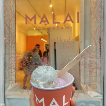 Malai - Cardamom pistachio and Rose with Cinnamon Roasted Almonds - A hand holds a cup of two-flavor ice cream with a Malai-branded wooden spoon in front of the Malai ice cream shop window in Brooklyn, NY. - Ice Cream & Frozen Yogurt Near Me - Brooklyn, New York