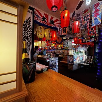 Dragon Sushi - The image shows the interior of a Japanese-themed restaurant decorated with red paper lanterns hanging from the ceiling, colorful banners with Japanese writing, and anime posters on the walls. The space includes a bar area with bottles and a seating area with booths and chairs. The decor emphasizes traditional Japanese elements combined with modern anime culture, creating a lively ambiance. - Sushi Bars Near Me - Albuquerque, New Mexico