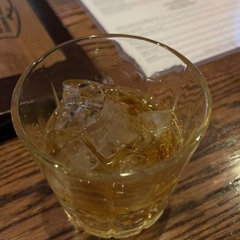 Spears Bourbon Burgers Beer - Ice?! - A clear glass contains a golden-colored whiskey with several ice cubes floating on top. The glass is positioned on a wooden table, and a menu or paper is partially visible in the background, suggesting a dining or bar setting. - New American Near Me - Wheeling, Illinois