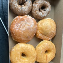 Half dozen assorted donuts. - Six donuts are arranged in a cardboard box, featuring a variety of types. Two donuts at the top are chocolate with chocolate chips and a light glaze. The middle row has a powdered sugar donut and a glazed donut with a shiny surface. The bottom row contains two glazed donuts with a smooth, shiny coating. The donuts are placed on a brown paper-lined box, likely from a bakery or donut shop.