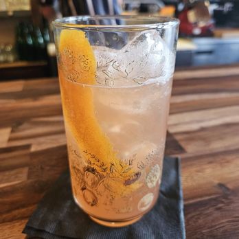 Broderick - After All Spritz - A transparent glass contains a clear, carbonated beverage with ice cubes and a lemon wedge. The glass is decorated with a floral pattern and is set on a black napkin on a wooden bar counter. The background shows a blurred bar setting with shelves and bottles. - American Near Me - Denver, Colorado