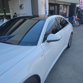 Wrapros - Audi A6 - A white sedan is parked on a paved surface outside a business that appears to sell tires and motorcycles. The shop has a display of motorcycle tires and a motorcycle visible near the entrance. Other vehicles are parked nearby, and a person is seen in the background near the building. The scene is outdoors with clear lighting. - Vehicle Wraps Near Me - Montclair, California