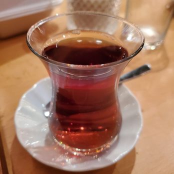 Meat Point - Turkish tea - A clear, tulip-shaped glass of Turkish tea filled with dark, brewed tea is placed on a white saucer. The glass is on a wooden table, with two other glasses visible in the background, one of which appears to contain water. The setting suggests a casual dining or tea-drinking environment. - Turkish Near Me - Toronto, Ontario