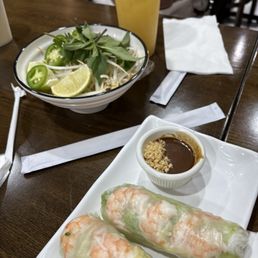 Summer Rolls - The image shows a wooden table with a plate of two shrimp spring rolls wrapped in rice paper, accompanied by a small bowl of dipping sauce topped with crushed peanuts. A bowl of salad containing bean sprouts, lime wedge, and sliced jalapeños is also present. A tall glass of yellow beverage, possibly juice or iced tea, is placed behind the salad. Napkins and utensils are arranged on the table, and a person wearing a white shirt is partially visible in the background.