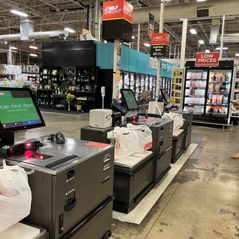 H-E-B - Self checkout stands for 10 items or less - The image shows a row of self-checkout stations in a grocery store, each equipped with digital screens, barcode scanners, and card payment terminals. Shopping bags are placed on the counters, and the area is set on a concrete floor. In the background, store signage indicates promotions and product sections, including refrigerated displays with various items. The store ceiling features exposed pipes and lighting fixtures, creating an industrial look. - Grocery Near Me - Waco, Texas