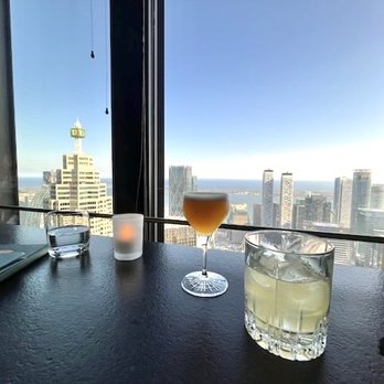 Canoe - Drinks with a view - A table with three drinks and a notebook is set against a large window overlooking a city skyline with tall buildings. - Canadian (New) Near Me - Toronto, Ontario