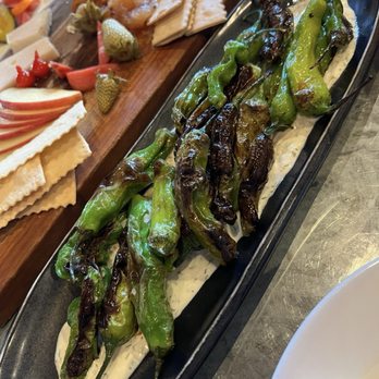 Revival Bar & Kitchen - SHISHITO PEPPERS - $13 - House-made crème fraiche ranch. - Charred shishito peppers are presented on a black platter with a creamy sauce, accompanied by a cheese and cracker platter in the background. - New American Near Me - Berkeley, California