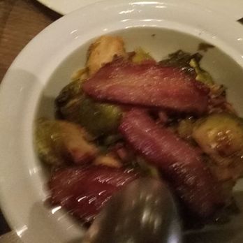 Mooo.... Beacon Hill - Brussel Sprouts - A white bowl contains Brussels sprouts topped with strips of cooked bacon, placed on a wooden table. - Steakhouses Near Me - Boston, Massachusetts