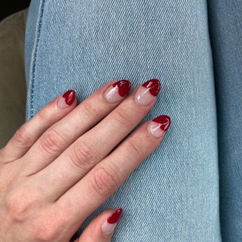 Elite Nails - A photograph shows a hand with neatly manicured nails, painted with a red and nude design, resting on light blue denim jeans. The nails are shaped in an almond style, and the background includes the fabric of the jeans, indicating a casual setting. - Nail Salons Near Me - Dubuque, Iowa